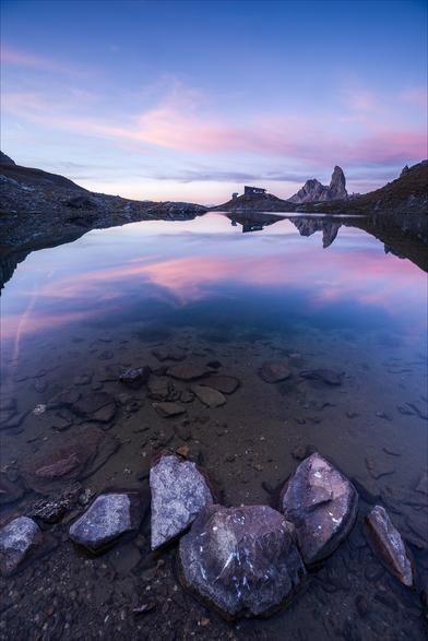 Photo d'un paysage de montagne au lever du jour, au format portrait.
Au premier plan, des cailloux baignent dans un lac, probablement près du bord vu la faible profondeur.
Tout au bout du lac se dressent un refuge et une montagne en forme de canine. Le ciel est bleu avec de fin nuages roses qui se reflètent dans le lac.
-
Portrait format photo of a mountain landscape at sunrise.
In the foreground, pebbles bathe in a lake, probably close to the shore given the shallowness of the water.
At the far end of the lake stands a refuge and a canine-shaped mountain. The sky is blue, with fine pink clouds reflected in the lake.