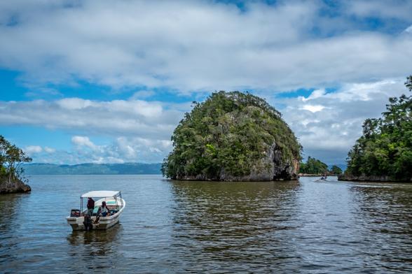 A small boat is anchored in calm waters, surrounded by lush green islands and mountains under a partly cloudy sky.