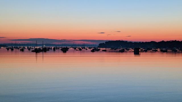 Historical Documentation in the form of a photograph of a rising sun over the ocean bay at the Historic Pioneer Village in Forest River Park, Salem, MA. The sky is a mix of orange, pink, and blue hues, with the sun rising in the background. The water is calm and reflects the colors of the sky. In the foreground, there are several boats docked in the water, with their sails dreaming of billowing in the wind. On the right side of the image, there is a small island with trees and mountains in the distance. The overall mood of the photograph is peaceful and serene.