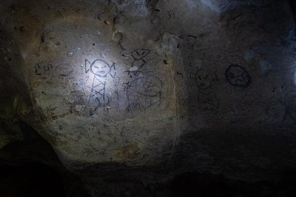The image shows ancient rock art depicting various figures, illuminated by a flashlight. The carvings include humanoid shapes and other abstract designs on a textured stone surface.
