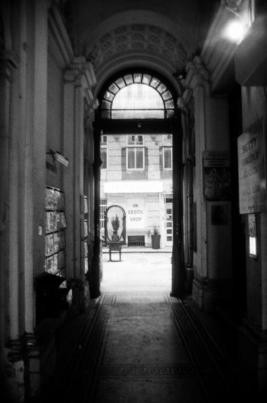 Black and white film photo of a passage leading into a courtyard. The doorway is arched with low lighting in the passage. In the courtyard there is an erotic shop window front. The courtyard is in bright sunshine.
Taken in Budapest, Hungary February 2025.