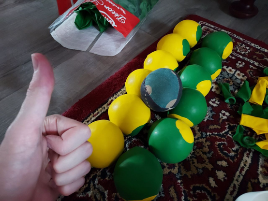 My thumb in front of 12 newly crafted juggling balls, half yellow and half green, on an old-fashioned carpet.