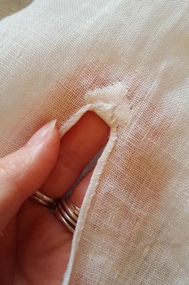 A very close up photo of the very tiny little hem hand sewn onto the slit on what is to become a blouse sleeve. The turn is some millimetres wide, and there is a tiny little square of folded up fabric hand sewn onto the slit end, where there will be some strain. The square has been flipped on the diagonal, and one corner has been folded over from the right side to the back. The fabric is a very delicate linen with some thread variation, sheer enough to see the shadow and colour of Sini's pink fingertips through it, as she is holding up the tiny detail for the camera.