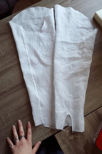 Two slightly strange shaped sleeves, of the same fine white linen, laid out on a beige wood grain desk. The sleeve seam is not at the very centre of the scoop that attaches to the armpit, but a bit to the front. This means that in a very soft and fine fabric the rigidity and added tension of the sleeve seam will not drag at the curve of arm and elbow, but naturally fall below the arm. The little sleeve slits are hanging open, not being attached to cuffs yet, with their little reinforcement squares. The sleeve seam has been done in a narrow flat felled hem, slightly wobbly.