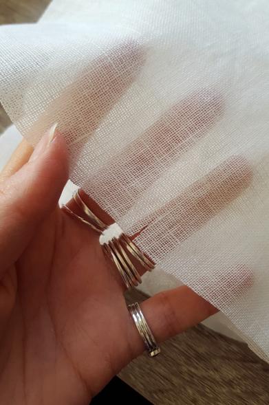 A photo showing how sheer and light the linen fabric is. White and shimmery, it shows through the details of Sini's hand behind it. There is variation in the thread weight and thickness, but at a glance and without focusing on it overmuch it looks incredibly nice and finely made. This is almost peak linen, these days. Sini's myriad silver rings glitter prettily, also.