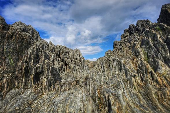 A low-angle shot looking up at jagged, grey-brown beach rocks under a blue sky with scattered white clouds. The nature of the rocks and the angle of the photo makes them appear like huge cliffs.