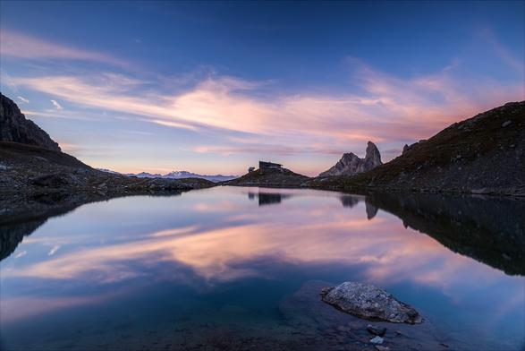 Photo d'un paysage de montagne au lever du soleil.
Un caillou baigne dans un lac, on devine à la profondeur que la berge est toute proche. 
Au bout du lac se trouve un refuge sur un petit promontoire encadré de part et d'autre par deux collines, juste à sa droite se dresse une montagne en forme de canine. Juste à gauche du refuge, on distingue très loin à l'horizon des montagnes dont certaines sont enneigées.
Le ciel est bleu, traversé de fins nuages rose pâle qui se reflètent dans le lac.
-
Photo of a mountain landscape at sunrise.
A pebble lies in a lake, and you can tell by its depth that the shore is nearby. 
At the end of the lake stands a refuge on a small promontory framed on either side by two hills, with a canine-shaped mountain just to its right. Just to the left of the hut, you can make out mountains in the distance, some of them snow-capped.
The sky is blue, with fine pale pink clouds reflected in the lake.