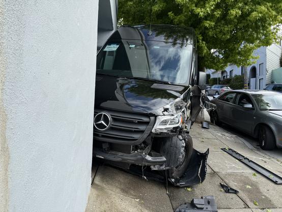 A black Mercedes-Benz Sprinter van has crashed into the corner of a light blue building. The van’s front right side is heavily damaged, with the bumper detached, the wheel pushed inward at an angle, and pieces of debris scattered on the sidewalk. A tree with green leaves partially covers the right side of the van. A gray sedan is parked adjacent to the van on the right, with minor visible damage to its rear side. The sidewalk appears slightly tilted, and there’s a parking sign in the background. A sticker inside the windshield of the van reads “Glen Park.”