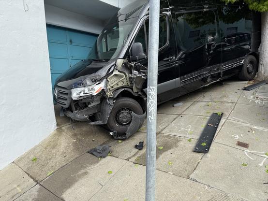 The same black Mercedes-Benz Sprinter van is shown from a wider angle. It has mounted the sidewalk and collided with the corner of a light blue residential building, partially blocking a teal garage door. The front right side is severely crushed, exposing the wheel well and suspension. The metal stop sign post in the foreground is leaning, likely impacted by the van. The side panel of the van is dented and scraped along its length. Tree leaves obscure part of the upper vehicle, and the van’s rear end is very close to a parked gray sedan. A man is standing on the sidewalk near the back of the van, looking toward the damage.