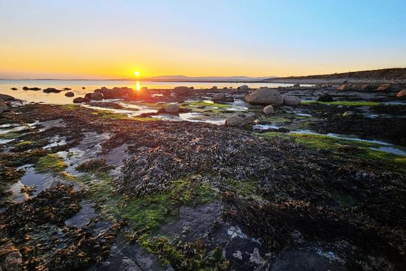 A vibrant sunset over a rocky beach in Ireland. The sun is setting, casting a golden glow across the sky and reflecting on the calm water. The rocky shore adds texture to the scene, creating a peaceful and serene coastal image.