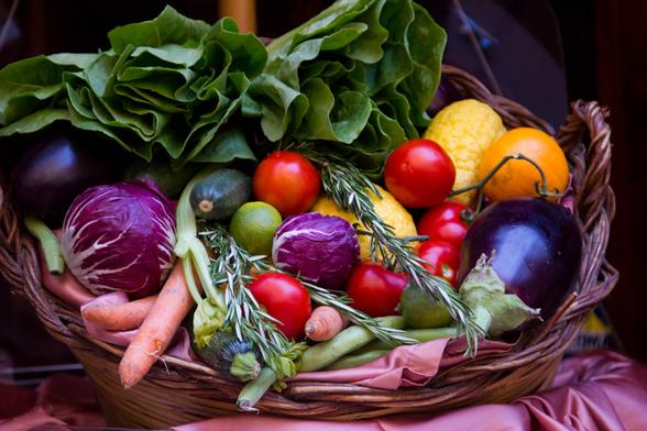 A large oval wicker basket that takes up the entire photo, filled with colorful, artfully arranged vegetables: tomatoes, red cabbage, lime, pumpkin, eggplant, carrots, etc. At the top is a row of green lettuce leaves.