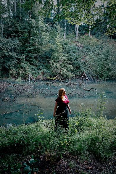 Rothaarige Frau in einem schwarzen Rock und einem roten Oberteil, die vor einem Waldsee steht, in den Äste ragen.