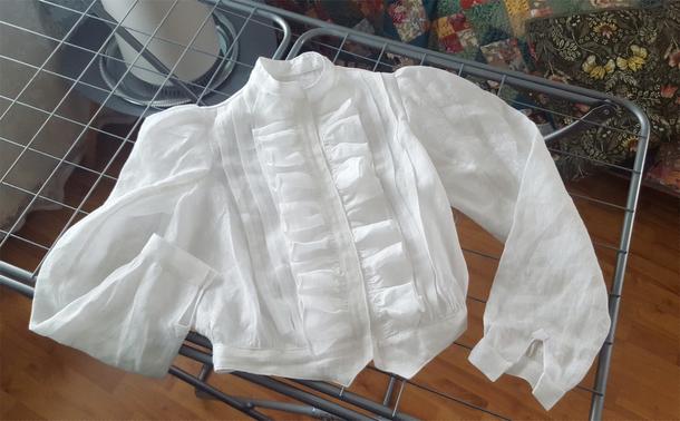A top down view of a mostly finished looking white linen blouse on the convenient surface of a metal wire rack for clothes. The sleeves have been folded a bit to fit them into frame, like the shirt is clutching at its pearls. The nearly insubstantial, thin fabric flows down from the shoulder in puffy sleeves that narrow down to a simple cuff, there's three decorative tucks running down the front, and a rather fancy and full ruffle both sides of the button placket. A narrow standing collar and a shaped waistband with a sort of dovetail notch at the front complete the style, which looks fairly historical but also just on the edge of something you recognise as modern, being Edwardian. An air purifier cylinder, some quilts and warm toned parquet in the background.