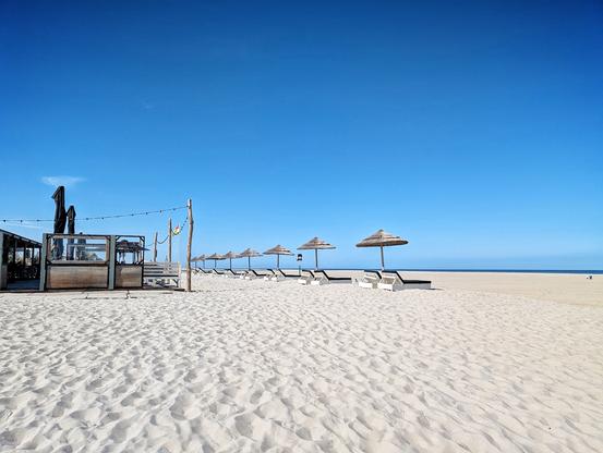 The image captures a serene and sunny day at the beach. The sky is a brilliant, cloudless blue, stretching out to the horizon. The sand is light-colored and appears soft, showing patterns of footprints and possibly the gentle movement of wind.
Along the edge where the sand meets what looks like a beachside establishment, there are several wooden structures. To the left, there's a more enclosed building with some signage and what appear to be string lights hanging above. Moving to the right, there are more open, wooden structures with benches.
Further along the beach, a neat row of thatched umbrellas provides shade over empty beach chairs. The umbrellas are evenly spaced, suggesting a calm and organized setting. In the far distance, a hint of the ocean can be seen as a thin, dark blue line meeting the lighter horizon. A few small figures are visible in the distance on the sand, emphasizing the vastness of the beach.
The overall impression is one of tranquility and relaxation on a beautiful day by the sea.