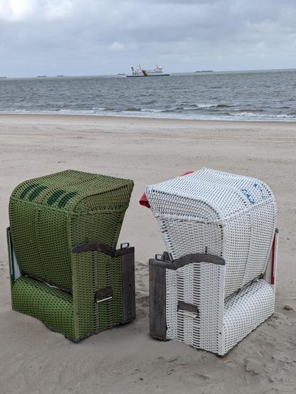 Ein grüner und ein weisser Strandkorb im Vordergrund.
Auf dem aufgewühlten Meer ist am Horizont eine Fähre zu erkennen.