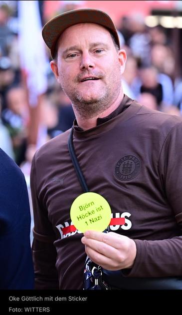 A man wearing a brown shirt with "FC St. Pauli" branding holds a bright yellow sticker that reads "Björn Höcke ist 1 Nazi." He has a casual appearance, topped with a colorful cap, and is surrounded by a blurred