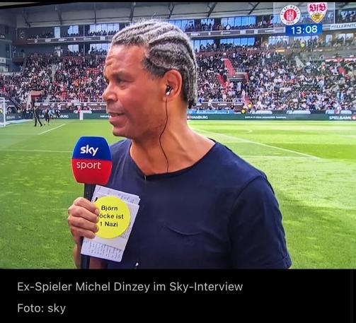 A television image of a sports interview featuring former player Michel Dinzey. He is holding a microphone with the "Sky Sport" logo and displaying a notepad with a message in German. The background shows a crowded stadium with football players on the field