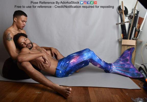 Two latino men, the first sitting on the ground holding the upper torso and arms of the second. He is looking down at the second, who is "passed out" (eyes closed) and wearing a mermaid tail.