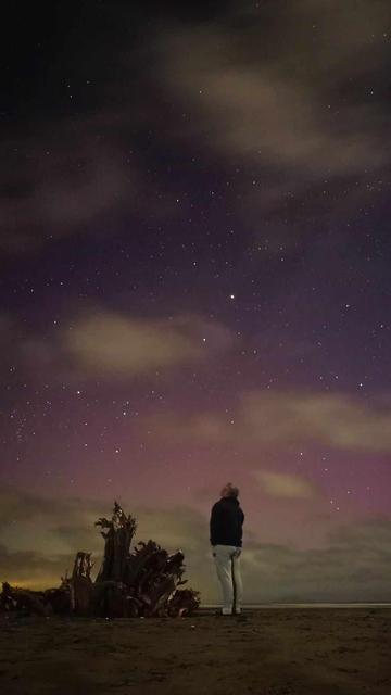Man on beach staring up at the cloudy aurora and stars. It's a majestical moment, a beautiful photograph.