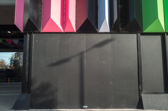 A shadow of a T-shaped light pole falls across the black wall of a building, which has some pink, black, white and green bits just above it. A tree and some sky is visible in the background over on the left.