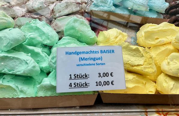 Baiser in vielen Farben hinter einer Fensterscheibe. Ein Schild zeigt die Preise und sagt: handgemachte Baiser, 1 Stück 3 Euro, 4 Stück 10 Euro. Die Farben der Baisers sind gelb, grün, blau und unklar (vielleicht mit etwas Schoko beigemischt)