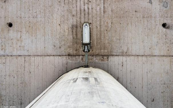 Blick nach oben an einer Betonsäule, an der eine Straßenlaterne befestigt ist. Darüber eine Betondecke. /
View upwards of a concrete column supporting a street lamp. Above, a concrete ceiling.