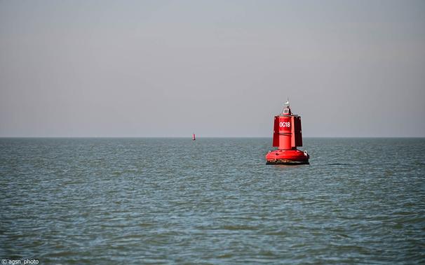 Blick vom Strand auf eine große rote Boje, die fest im Wasser verankert ist und die Schifffahrtsroute entlang der Küste markiert. /
View from the beach of a large red buoy that is permanently fixed in the water and marks the shipping route along the coast.