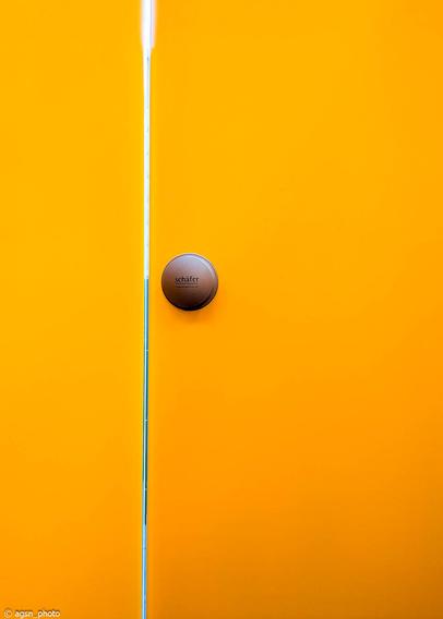 Ein runder silberner Türgriff an einer gelben Umkleidetür, links daneben ein schmaler Türspalt und eine gelbe Seitenwand. /
A round silver door handle on a yellow changing room door, to the left of it a narrow gap in the door and a yellow side wall.