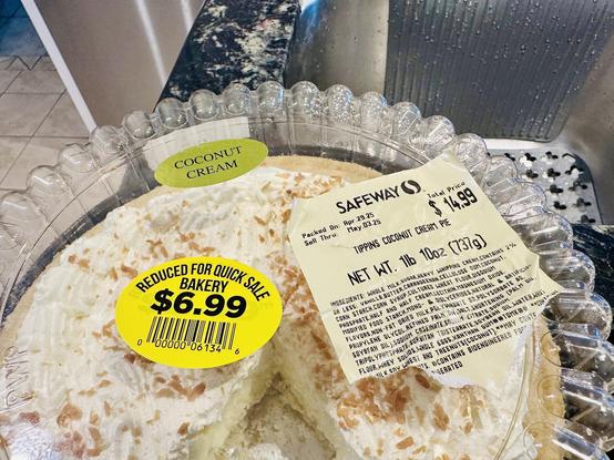 Top half of covered coconut cream pie from grocery, on counter by sink. The labels are on top of the hard clear plastic cover. Left one shows $6.99 reduced for quick sale and the other the ingredients and $14.99 regular price. One piece of pie center bottom is missing.