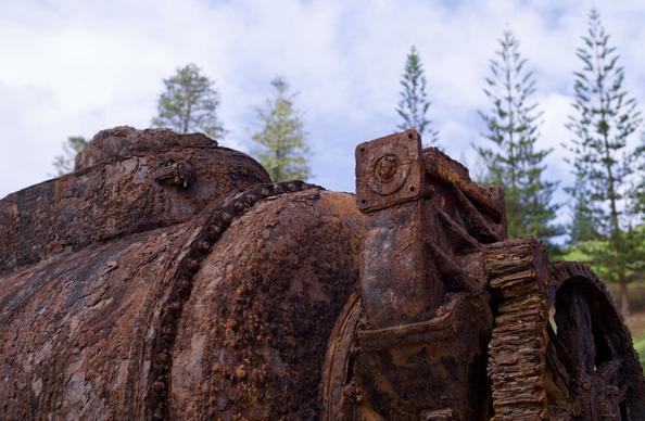 Photograph of rusted old engine sitting outside. The engine is a series of components bolted together. At one end is a large round tank, and the other is a large toothed gear wheel.