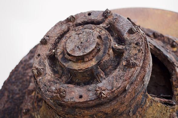 Photograph of rusted old engine sitting outside. The end of a large round tank is in the centre of the image. A series of concentric circular components are all decomposing with rust. Some bolts are still visible, but holes show where others would once have been.