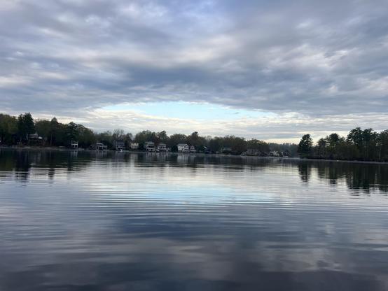 Lake with trees and trees in the back ground. A cloudy day reflected in the water. A circle of blue sky in the center of the clouds.