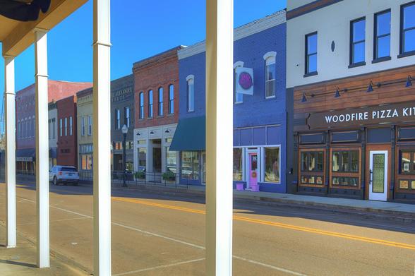 A vibrant small-town street is lined with colorful brick storefronts, displaying signs for local businesses, including a pizza kitchen.