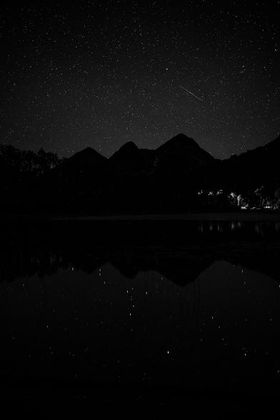 This image shows a night scene with a star-filled sky and a silhouette of mountains or hills.

There is a reflection of the sky and mountains on a body of water in the foreground, creating a mirror effect.
