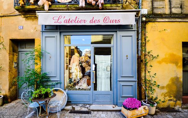 Eine kleine Boutique mit kleinem Schaufenster und schmaler Eingangstür. Die holzgetäfelte Fassade ist blau gestrichen.
Davor steht ein kleiner rostiger Gartentisch mit zwei weißen Metall-Gartenstühlen. Auf der rechten Seite dient ein alter Puppenwagen als Blumentopf. /
A small boutique with a small display window and a narrow entrance door. The wood-paneled exterior is painted blue.
In front of it is a small rusty garden table with two white metal garden chairs. On the right side, an old doll's pram serves as a flowerpot.