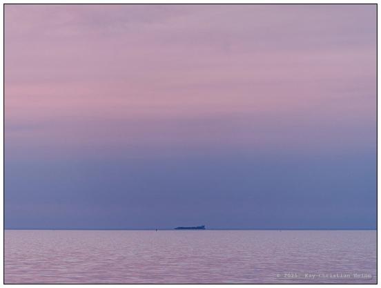 Ruhevolle Ostseeküste bei Hohenfelde im nördlichen Kreis Plön, die Horizontlinie verläuft im unteren Bildviertel, Wasser und Himmel sind einheitlich in sanft-pastelligen rosa-, violett- und Blautönen gefärbt und bilden so einen einheitlichen Raum, mittig ein Schiff am Horizont.