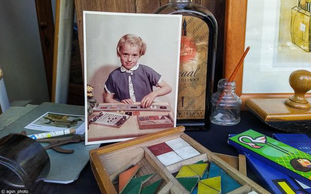 Im Schaufenster steht eine Holzkiste mit geometrischen Figuren, dahinter ein älteres Werbefoto von einem jungen Mädchen, das diese Figuren in ein Holzbrett einfügt. /
In the shop window there is a wooden box with geometric figures, behind it an older advertising photo of a young girl fitting these figures into a wooden board.