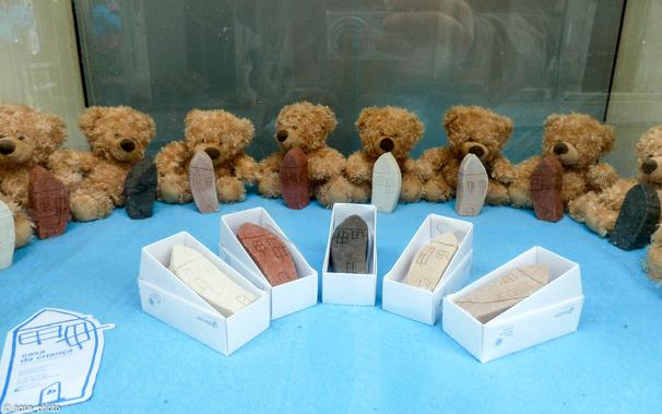 Im Hintergrund sitzen acht kleine Teddybären im Halbkreis, davor stehen kleine, leicht geschwungene Häuser aus Lehm mit spitzen Dächern und eingeritzen Fenstern und Türen. /
In the background, eight small teddy bears sit in a semicircle, in front of them are small, slightly curved houses made of clay with pointed roofs and carved windows and doors.