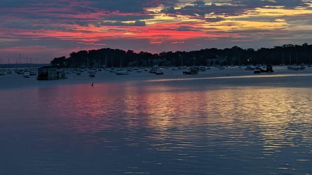 Historical Documentation in the form of a photograph of a rising sun over the ocean bay at the Historic Pioneer Village in Forest River Park, Salem, MA. The sky is filled with vibrant colors of pink, orange, and yellow, with the sun rising in the background behind the clouds. The water is calm and reflects the colors of the sky, creating a mirror-like effect. In the foreground, there are several boats and yachts docked in the harbor, with a few people walking along the shore. The harbor is surrounded by trees and buildings, and there is a small island in the distance. The overall mood of the image is peaceful and serene.