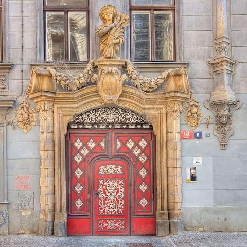A richly ornamented Art Nouveau doorway in Prague, featuring vibrant red double doors with intricate gold and black detailing. The entrance is framed by an elaborate stone archway adorned with floral swags, sculpted figures, and a central cartouche. Above the archway, a statue of a draped female figure holding a sheaf of wheat stands on a decorative pedestal. To the right of the doorway, blue plaques display the numbers "121" and "6." The building's facade is a muted gray, and the ground in front is paved with cobblestones.