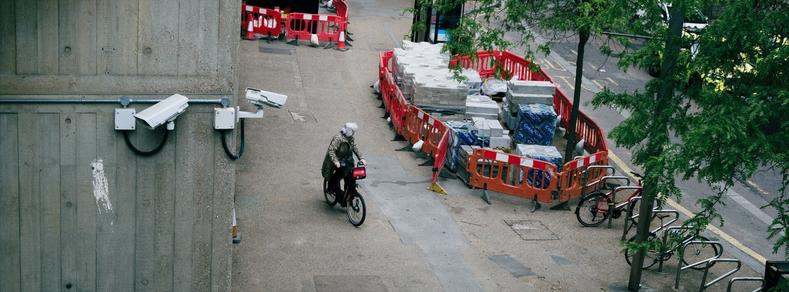 A person rides a red rental bike past a construction zone with stacked materials and orange barriers; security cameras and trees are visible on either side.