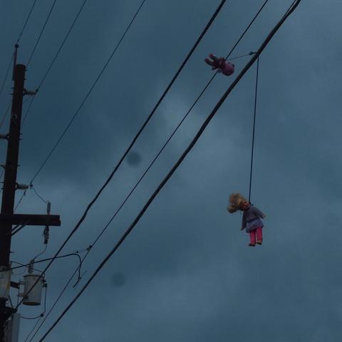 Photo of a doll hanging by its neck from a power line with a grey sky as the backdrop