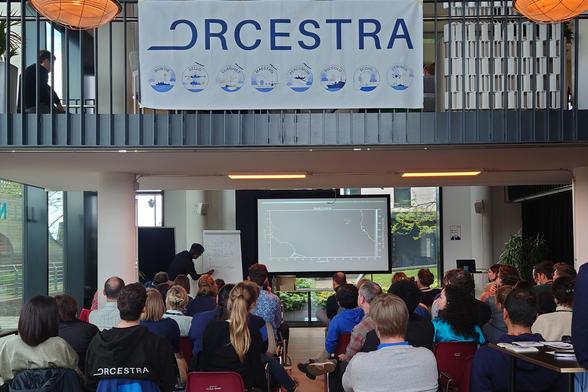 Participants seated in a modern conference room attend a workshop under a large banner that reads 'ORCESTRA'. The banner features illustrations of ships with names like Bowtie, Cello, Clarinets, Maestro, and others. A speaker is writing on a flip chart at the front, while a graph is projected onto a screen.