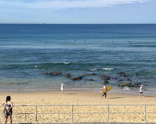 Looking over Bar Beach and out to sea. A woman in bathers and a towel is watching the beach scene from the terrace above the sand. A surfer is walking to the right with a bright yellow board under one arm. He's bookended by walkers wearing white tops and hat. Further out in the green-blue water, tiny black specks - surfers out waiting for a wave. Sunny morning. Happy, relaxed vibe.