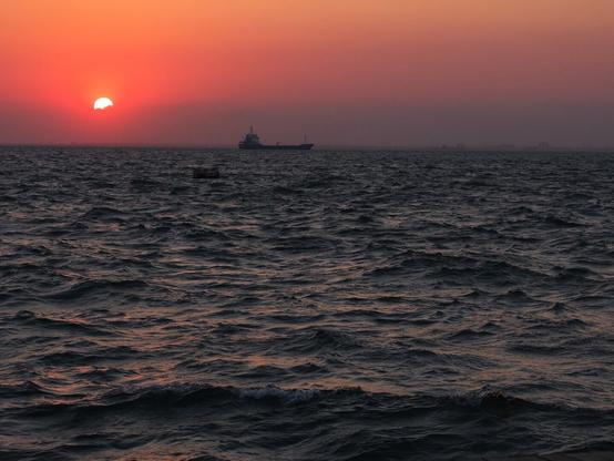 This image depicts a serene sunset over the ocean. The sun is partially visible on the horizon, casting a warm orange glow across the sky, while the water reflects the colors of the sunset. There is a ship in the distance, silhouetted against the vibrant backdrop. The waves in the foreground add texture to the scene, creating a peaceful and tranquil atmosphere.