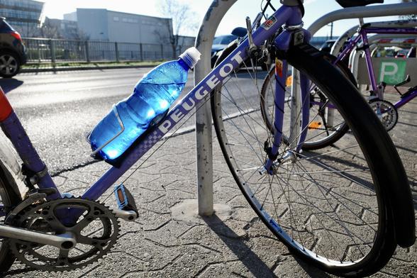 The color image shows a purple Peugeot bicycle locked to a bike rack on a sunlit street, with a bright blue plastic water bottle held in a cage on the bike’s frame and cars visible in the background.