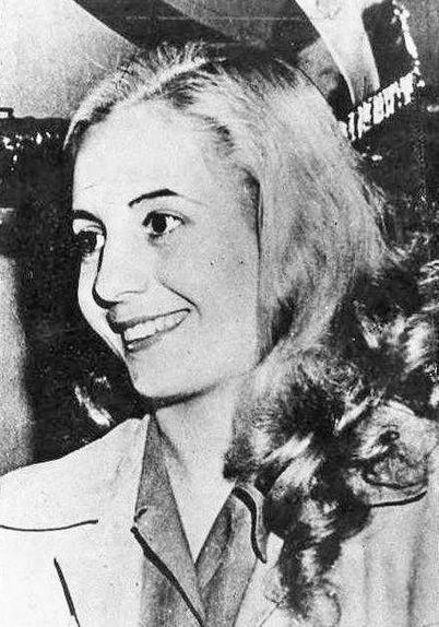Photo en noir et blanc d’Eva Perón, affichant un large sourire, vêtue d’un manteau clair avec des revers larges et des boutons visibles. Ses cheveux sont coiffés vers la gauche. L’arrière-plan est sombre et indistinct.