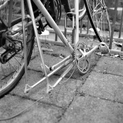 The black and white image shows the lower part of a bicycle, focusing on the crankset, pedals, and rear wheel, with the chain missing and the bike resting on a paved surface near a metal railing.