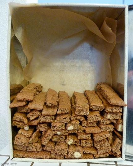 Blick in eine große Blechdose, die zur Hälfte mit in Streifen geschnittenem braunem Gebäck gefüllt ist. /
View into a large tin box half filled with brown pastries cut into strips.
