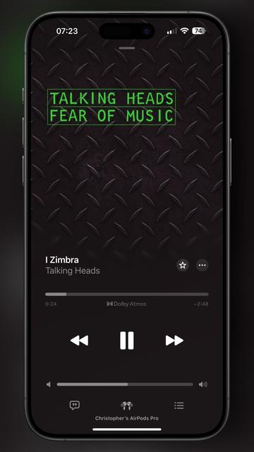 An image of a smartphone displaying the song "I Zimbra" by Talking Heads, from the album "Fear of Music." The background features a textured metal design, and the screen includes playback controls and airpod connection details.